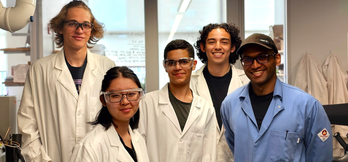 a group of diverse high school students wearing safety glasses and lab coats in a lab