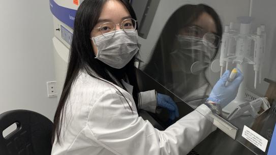 student wearing lab coat, mask and gloves working in a hood