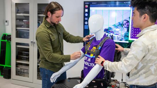 2 students in the lab putting a harness on a mannequin 
